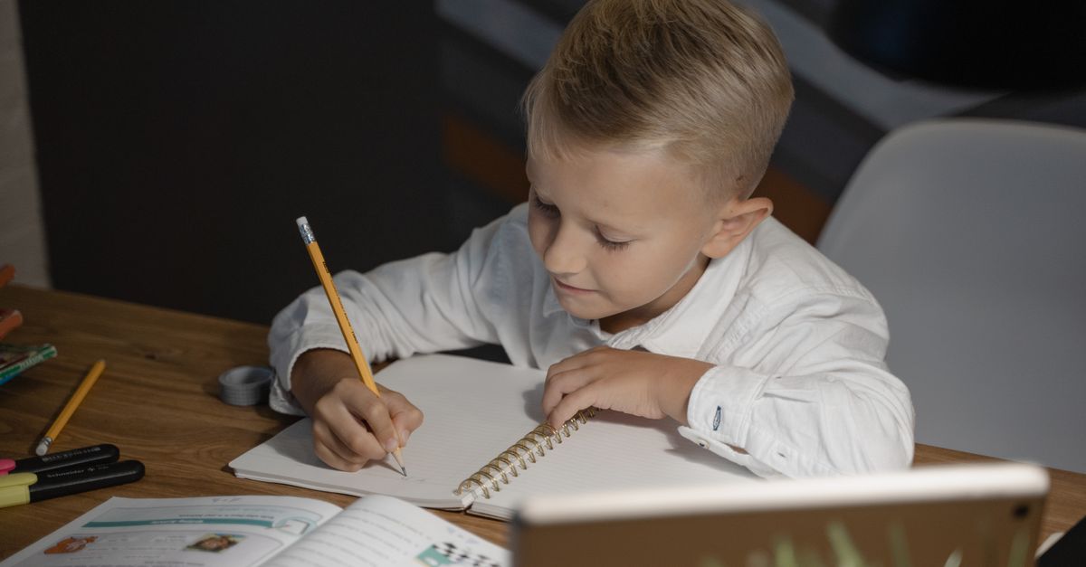 Menino escrevendo em um caderno, sentado à mesa, fazendo lição de casa