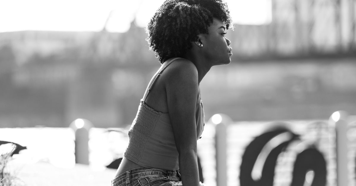 Retrato em preto e branco de uma mulher sentada à beira de um rio, pensativa e olhando para o horizonte